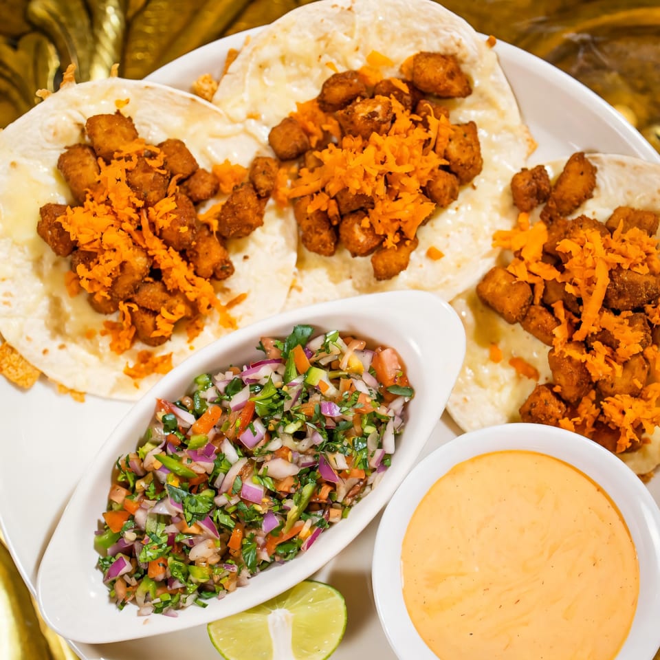 Three chicken tacos with shredded cheese, salsa, and a side of queso dip and lime.