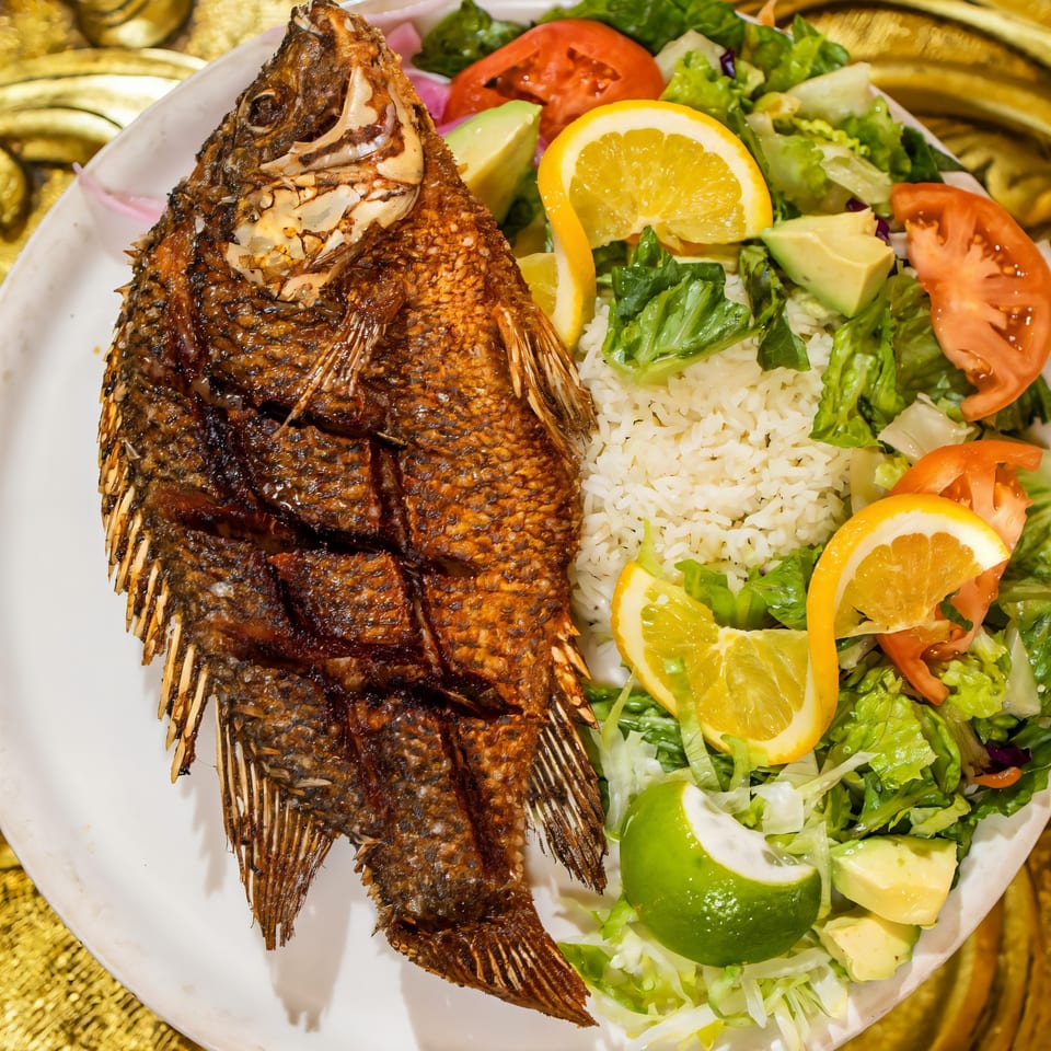 Crispy fried whole fish with rice, salad, avocado, tomatoes, and citrus slices on a white plate.
