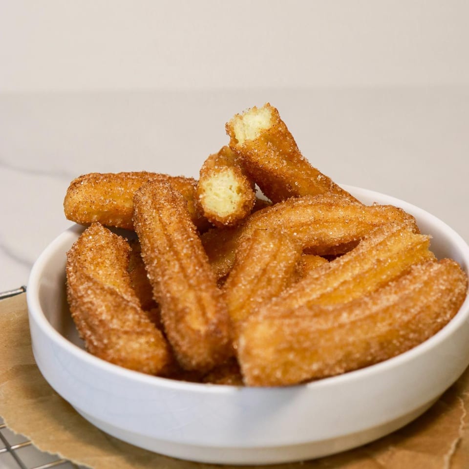 A white bowl filled with golden-brown churros, coated in cinnamon sugar, with one broken open.